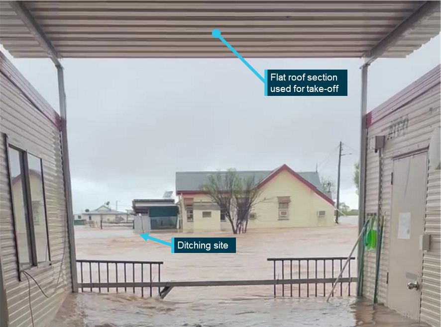 Video still showing a view from the flooded deck underneath the flat roof between two demountable buildings. The ditching site is visible across floodwater, the roof has supports on either side but not across.