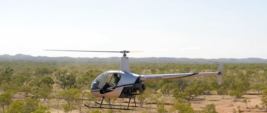 An R22 helicopter with doors removed flying over wooded terrain.