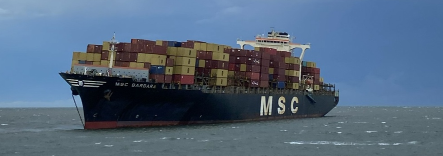 MSC Barbara aground in Koopa basin with a 5 degree heel to port. Its starboard anchor chain is visible running out across its bow and its port gangway is lowered halfway down.