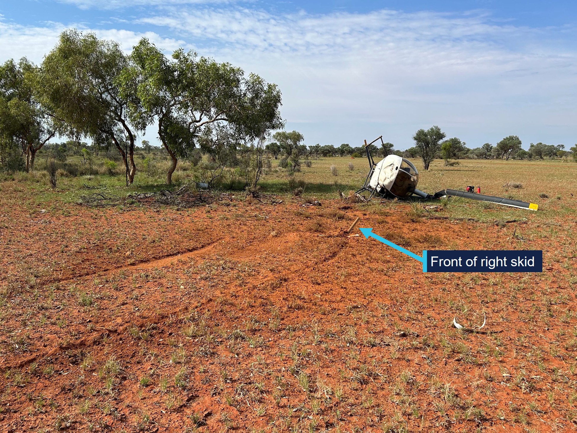 Image of flat, open ground with few trees. Two skid marks through the dirt in the foreground, leading to a helicopter on its side. The front of the right hand skid is sticking out of the ground at the end of the right skid mark.
