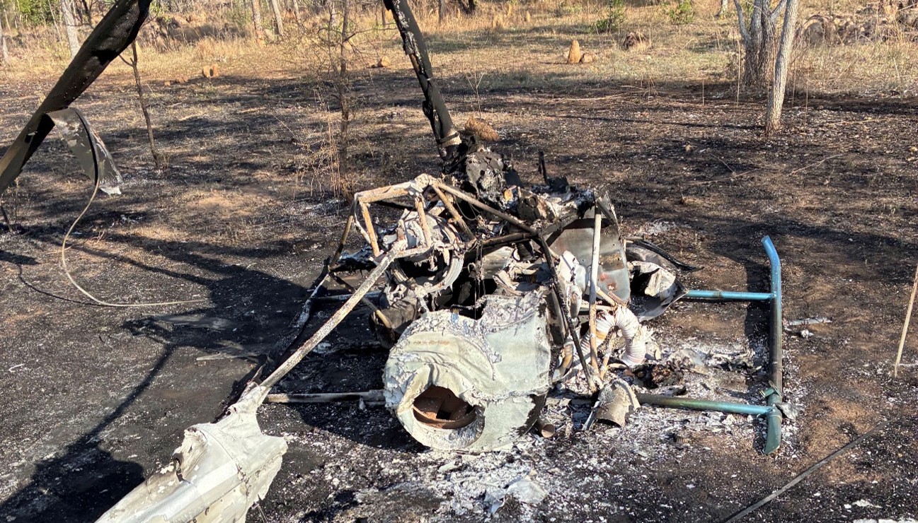 Completely burnt helicopter, pictured from the rear. Skids are spread flat, parallel to the ground. Area around the helicopter also burnt.