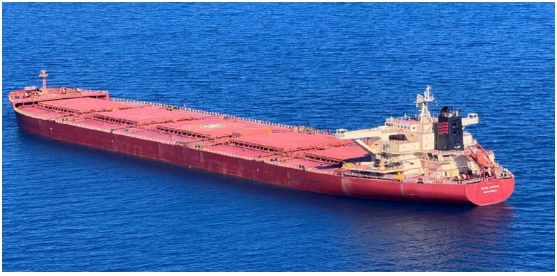 Photograph of RTM Weipa sailing across calm waters. The bulk carrier ship has a red hull, 5 cargo holds, and the off-white superstructure accommodation, funnel stack and free fall lifeboat at the stern. Cargo hold no. 3 features a heli-deck.