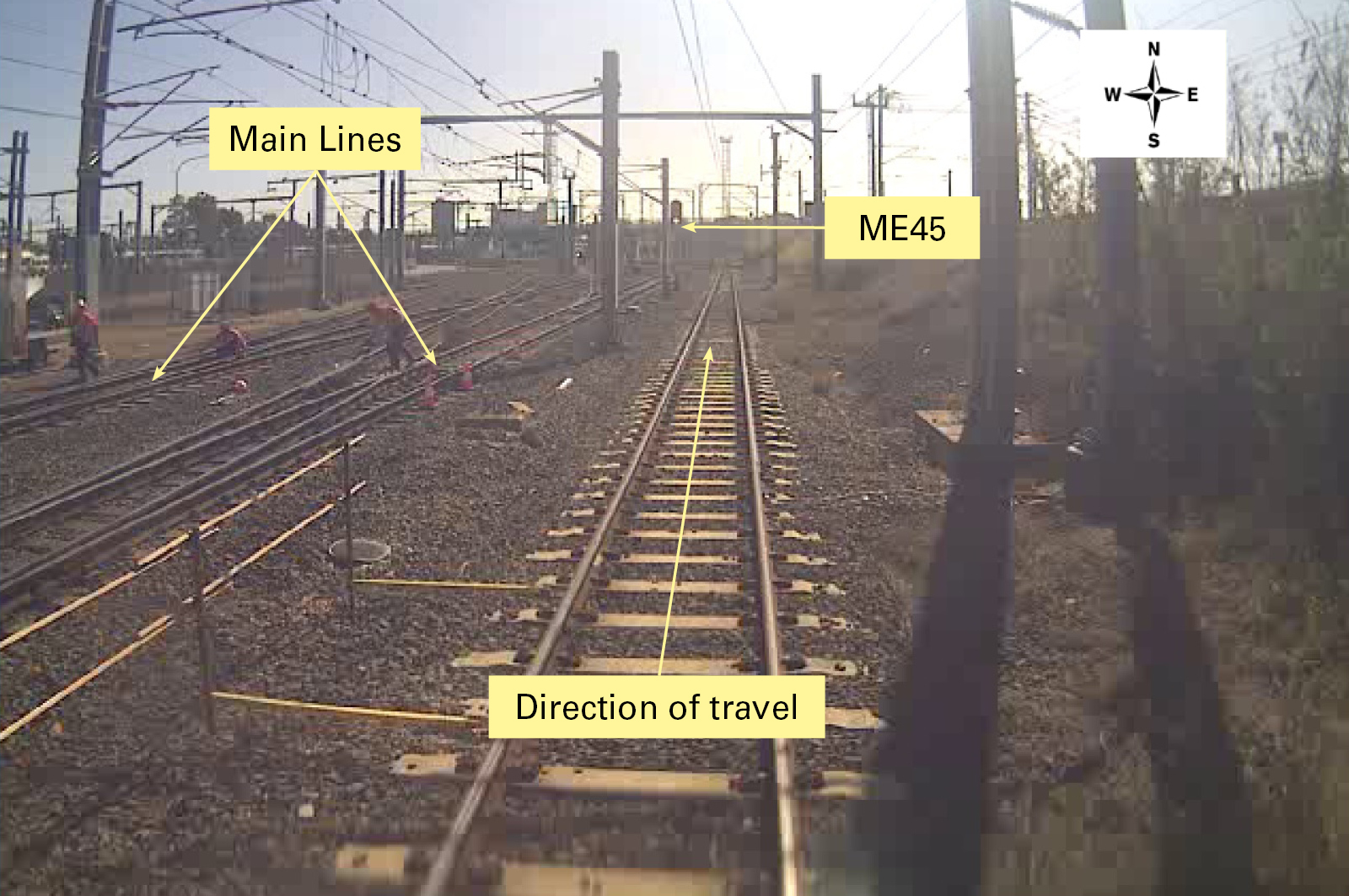 A still taken from CCTV footage from the driving compartment of train 1A21. Annotations show the main lines, the direction of travel, and the location of signal ME45.  It is likely that the driver’s attention was diverted to the workers on the left of the photo and away from signal ME45.<br />
Image supplied by Queensland Rail – Annotated by the ATSB.