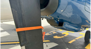 Image of an aircraft propeller blade and engine cowling. The propeller blade has an orange strap attached to it, and the engine cowling has pins attached.