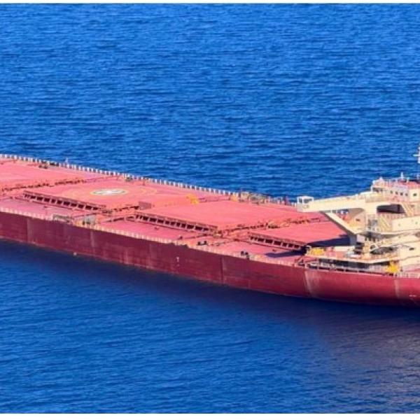 Photograph of RTM Weipa sailing across calm waters. The bulk carrier ship has a red hull, 5 cargo holds, and the off-white superstructure accommodation, funnel stack and free fall lifeboat at the stern. Cargo hold no. 3 features a heli-deck.