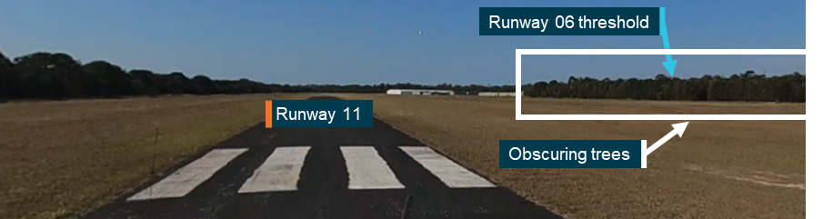 Photograph taken down runway 11 from the threshold. A highlighted line of trees obscured a portion of runway 06. The position of the obscured threshold of runway 06 is indicated with an arrow.