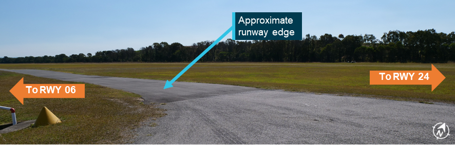 A photograph of the paved taxiway across runway 06/24. The approximate runway edge is shown with an arrow. No hold point markings are present.