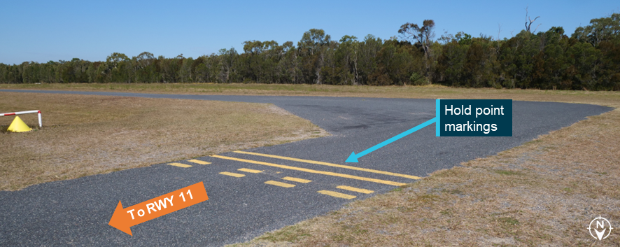 A photograph showing yellow hold point markings on a paved taxiway leading to runway 11. The markings consist of 2 unbroken lines across the taxiway followed by 2 dashed lines.