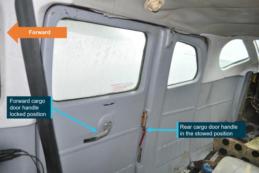 Internal image of a Cessna U206 G facing the closed cargo doors, the seats have been removed from this aircraft. Labels indicate the forward cargo door handle and obscured rear cargo door handle.