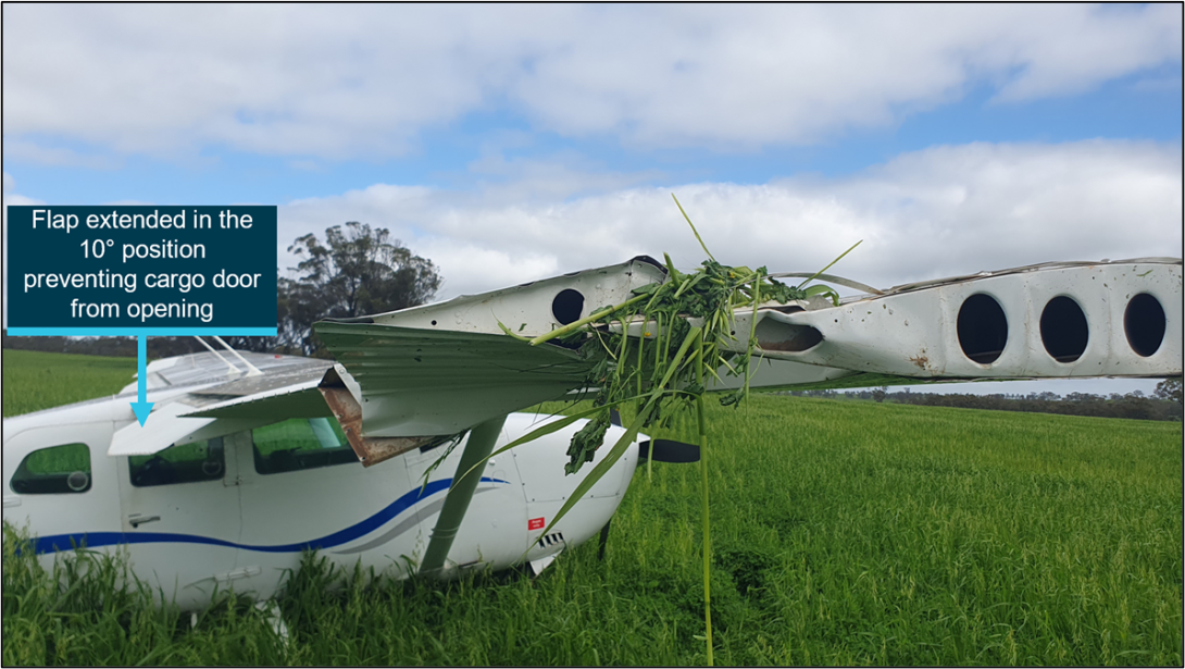 The front section of a Cessna 206 in a field with damage to the right wing tip. The flaps are extended in the 10 position and prevent the rear cargo door from opening.