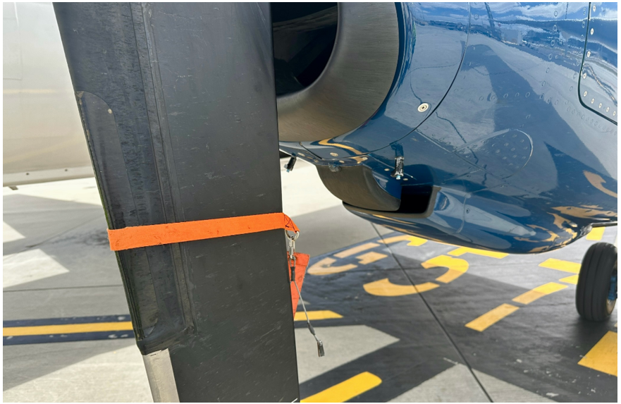 Image of an aircraft propeller blade and engine cowling. The propeller blade has an orange strap attached to it, and the engine cowling has pins attached.